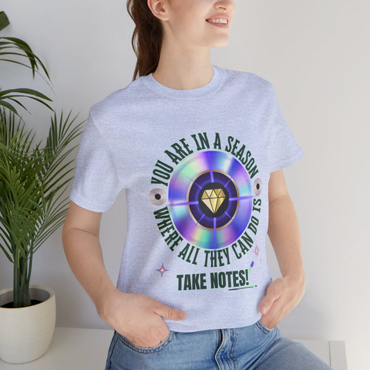 Person wearing a light blue t-shirt with a colorful graphic and text You are in a season where all they can do is take notes, standing indoors next to a plant