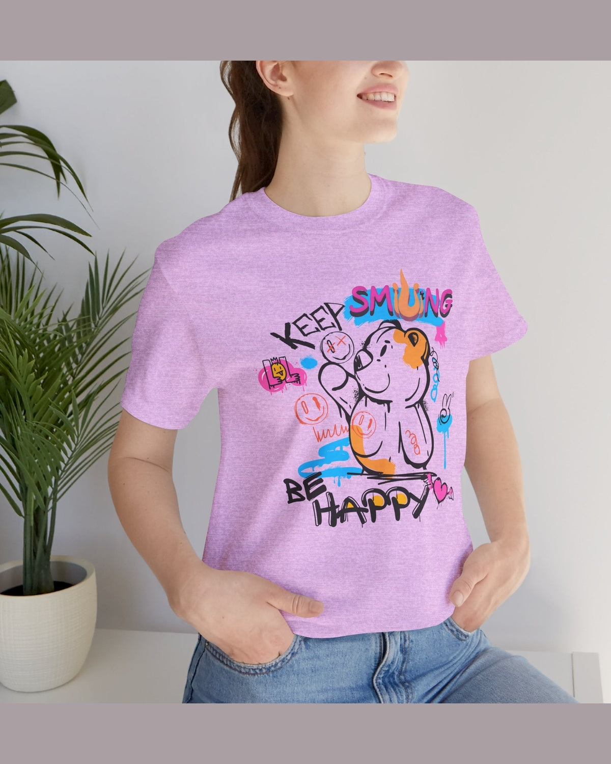 Person wearing a purple t-shirt with a colorful graphic and text Keep Smiling, Be happy, standing indoors next to a plant.