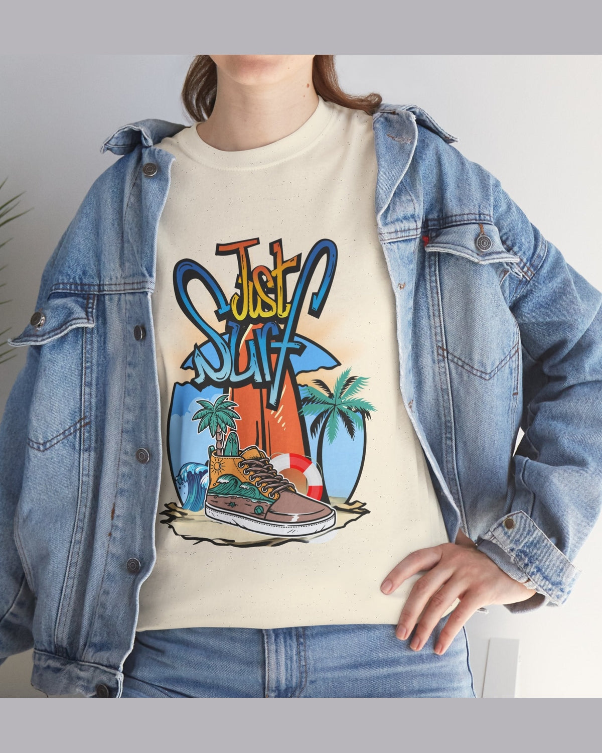 Person wearing a denim jacket over a cream t-shirt with a colorful graphic design with a text Just Surf