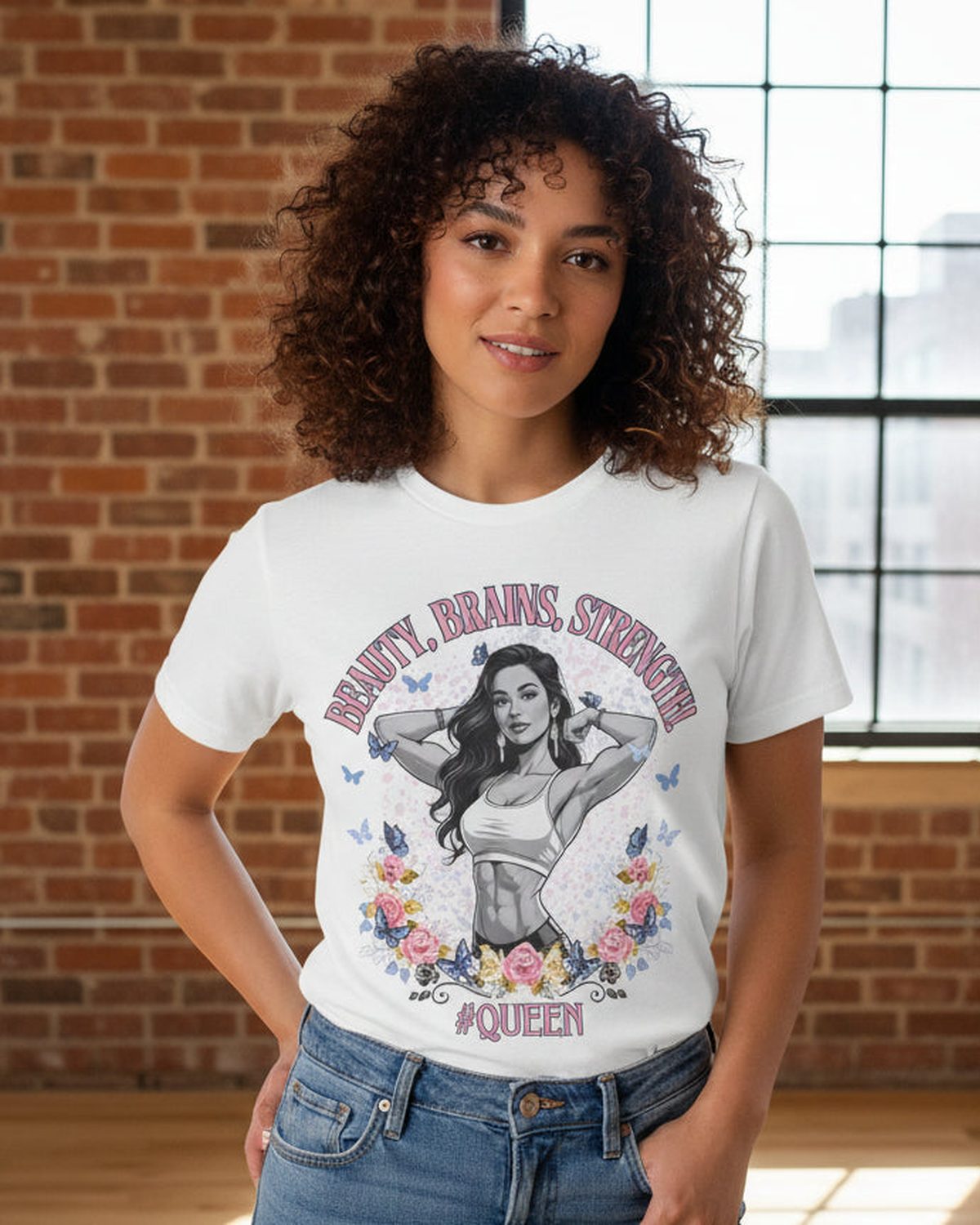 Woman wearing a white t-shirt with a graphic design of a fitness woman and a text Beauty, Brains, Strength #Queen' in front of a brick wall.