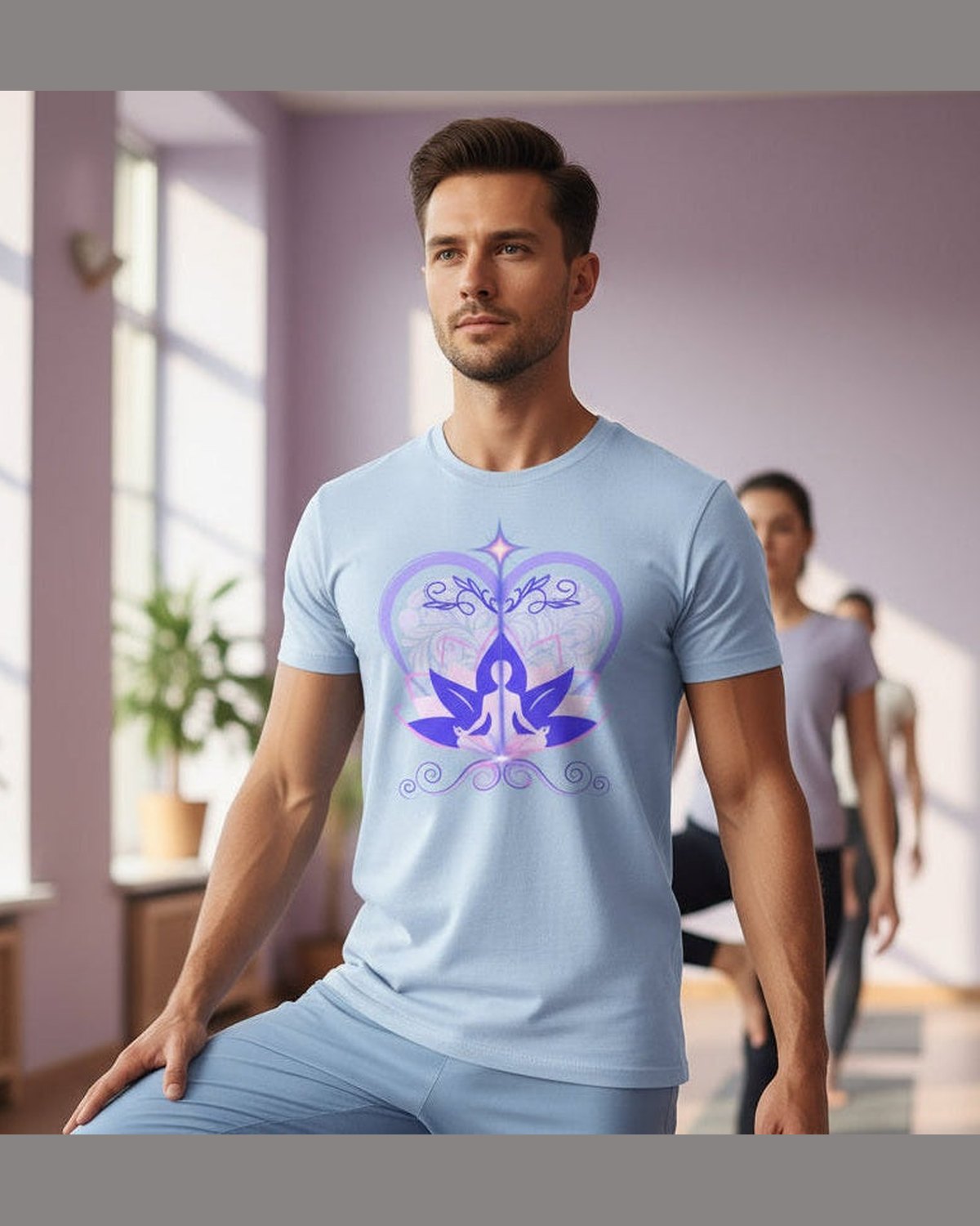 Man wearing a light blue t-shirt with a purple and pink design of a meditating figure, heart and lotus in a yoga room with other yogis and large windows and plants.