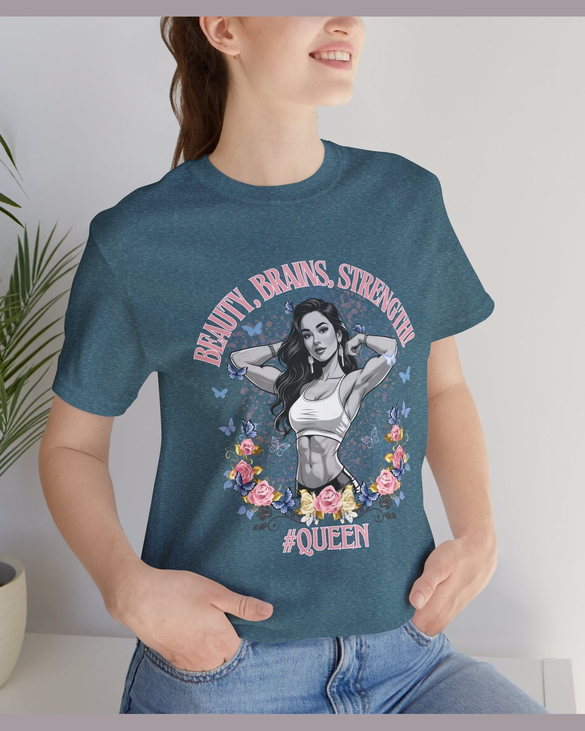 Person wearing a teal t-shirt with a graphic design of a fitness woman, flowers, butterflies and text 'Beauty, Brains, Strength #Queen' standing next to a plant.