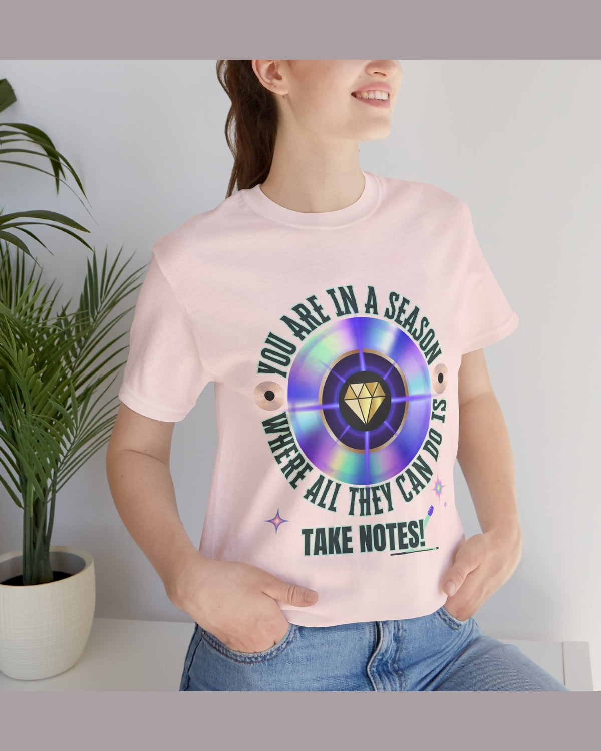 Person wearing a pink t-shirt with a colorful graphic and text 'You are in a season where all they can do is take notes', standing indoors next to a plant.