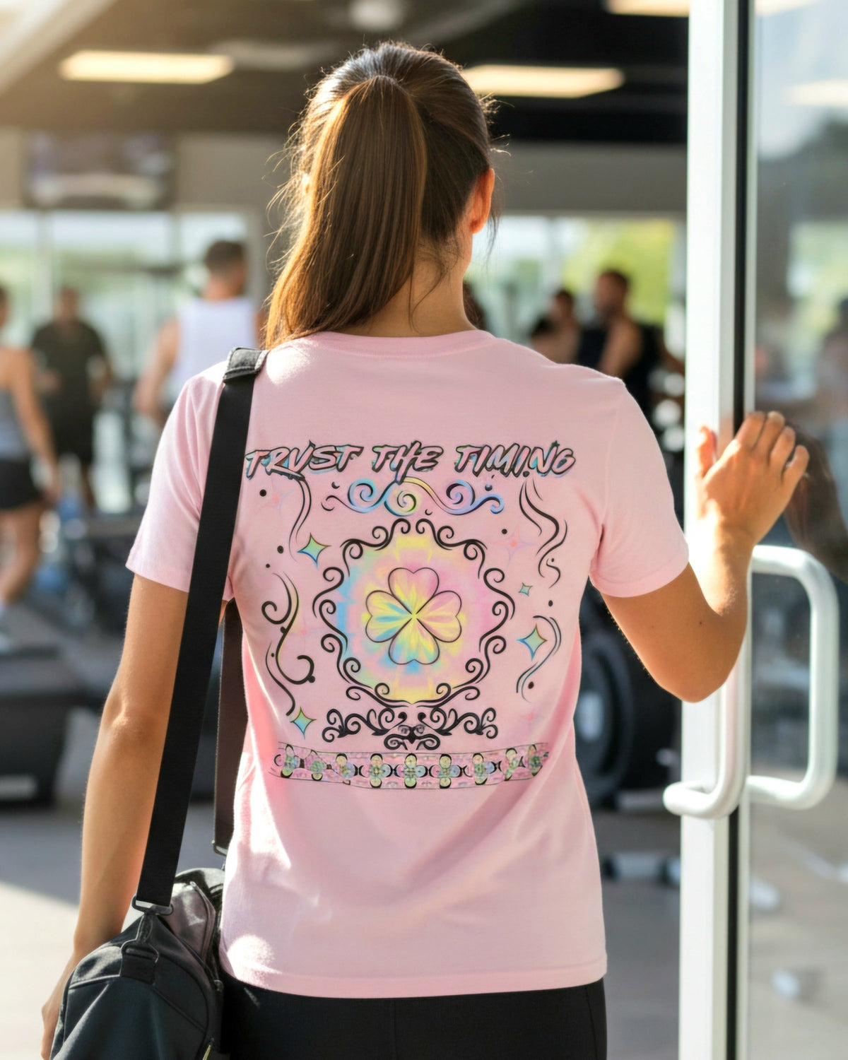 Woman is wearing a pink shirt walking into a gym the shirt has a vibrant four leaf clover and a text trust the timing
