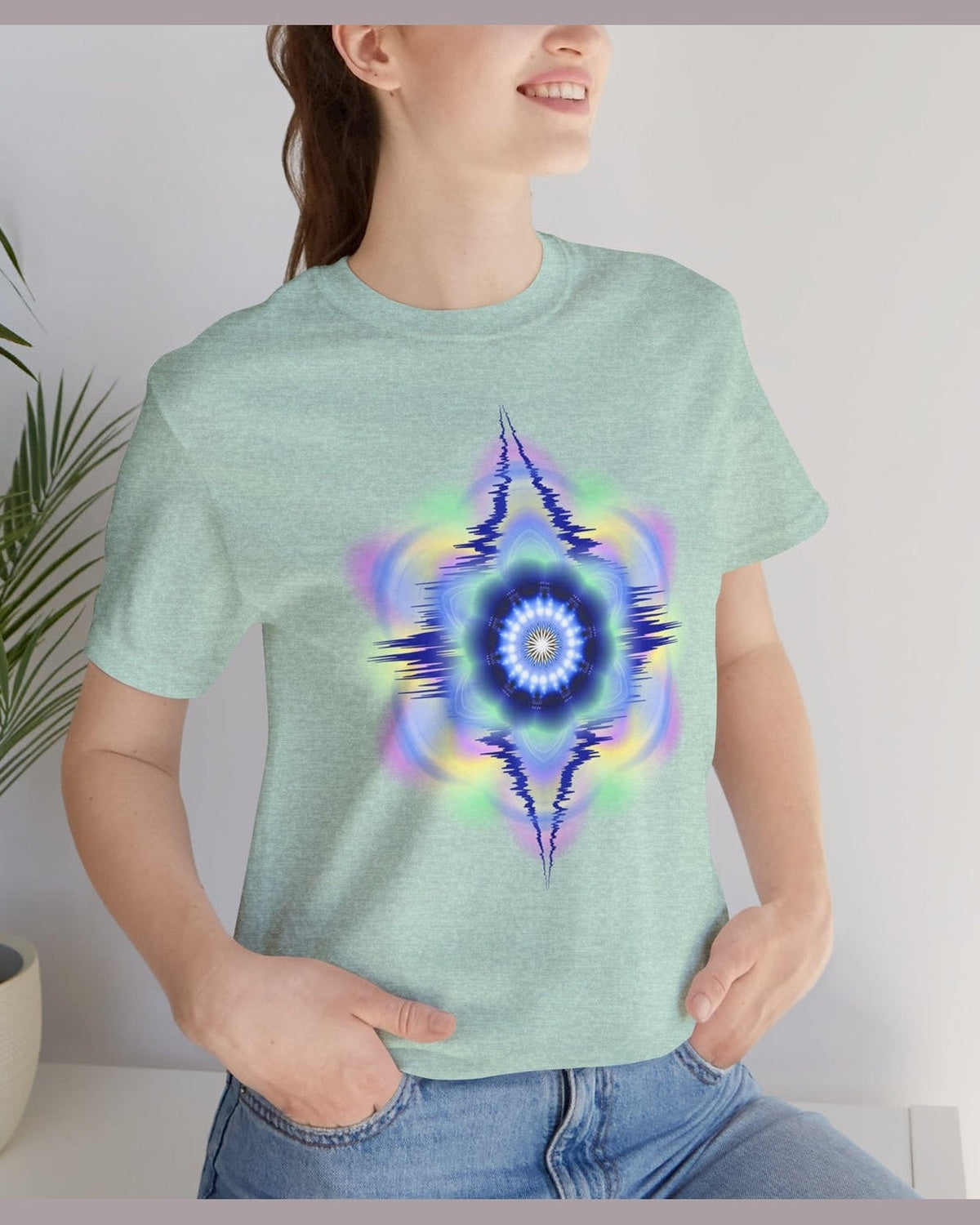Person wearing a light green t-shirt with a vibrant hues abstract design, standing indoors next to a plant.