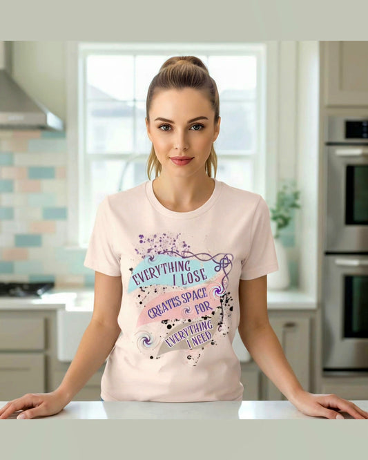 Woman wearing a pink t-shirt with a colorful graphic and text 'Everything i lose creates space for everything i need' in a kitchen.