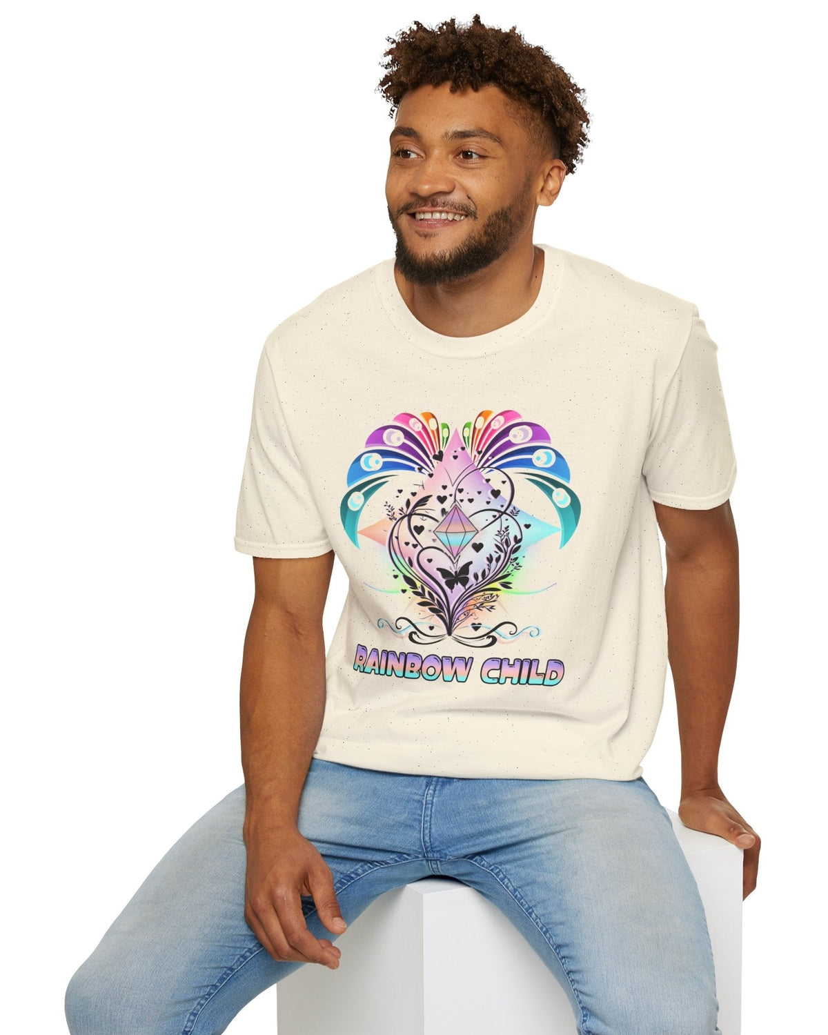 Man wearing a cream t-shirt with a colorful graphic and 'Rainbow Child' text, sitting on a white stool.