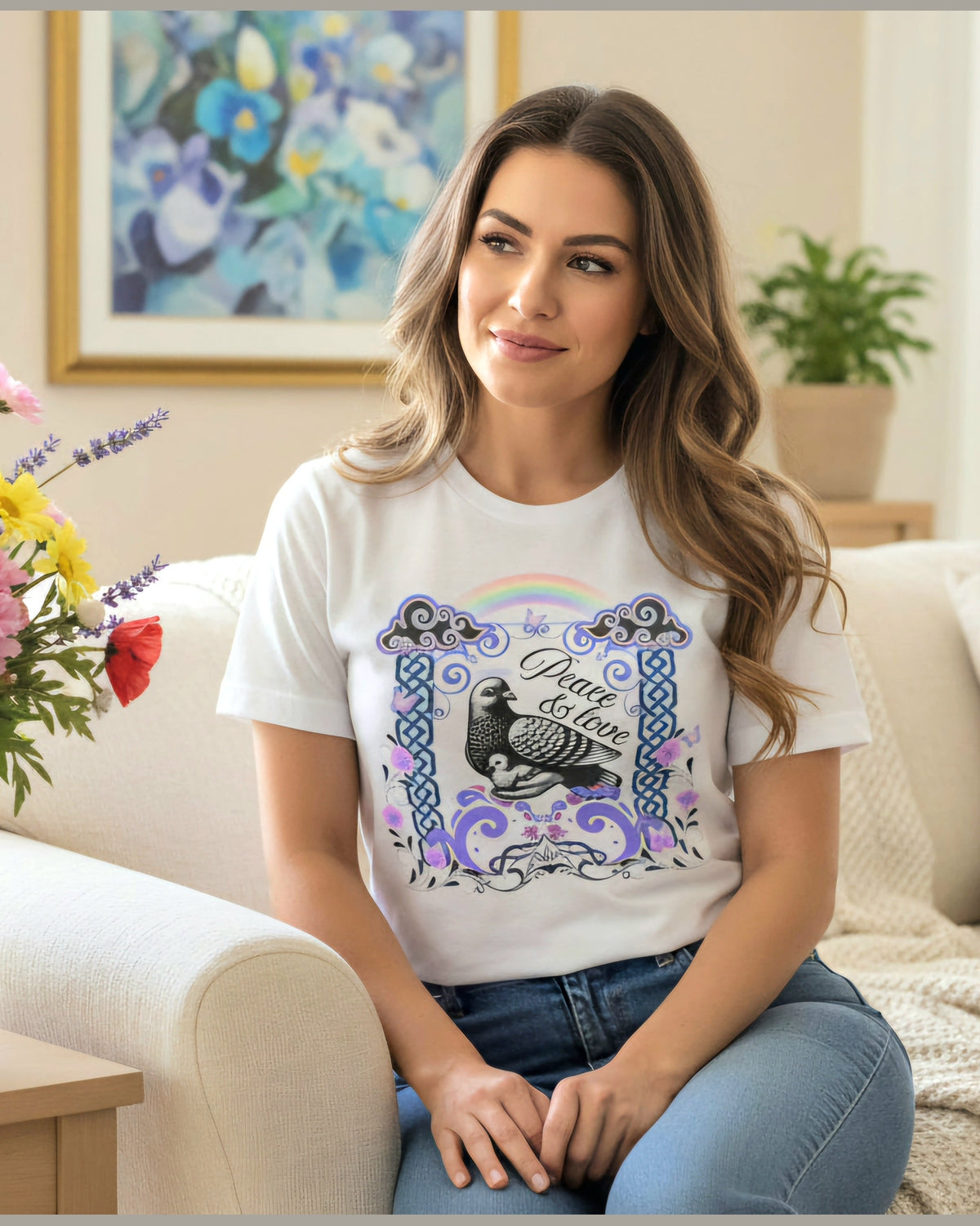 Woman sitting on a couch wearing a white t-shirt with a graphic Pigeon design and a text Peace & Love, surrounded by flowers and a painting.