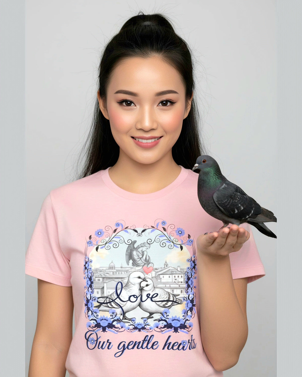 Woman wearing a pink t-shirt with a graphic design of pigeons in love and a text 'Love our gentle heart', holding a pigeon.