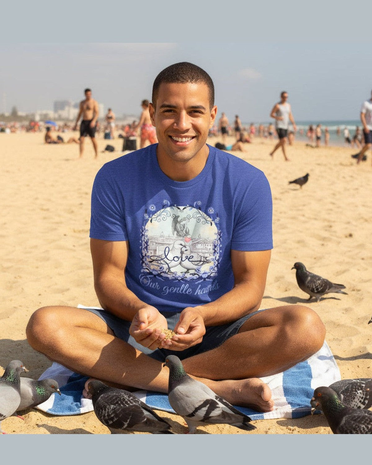 Man sitting on a beach with pigeons around, wearing a blue pigeon t-shirt.