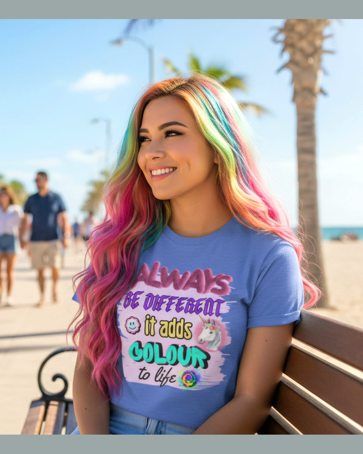 Person with colorful hair wearing a blue t-shirt with a motivational quote Always Be Different It adds colour to life sitting on a bench by a beach.