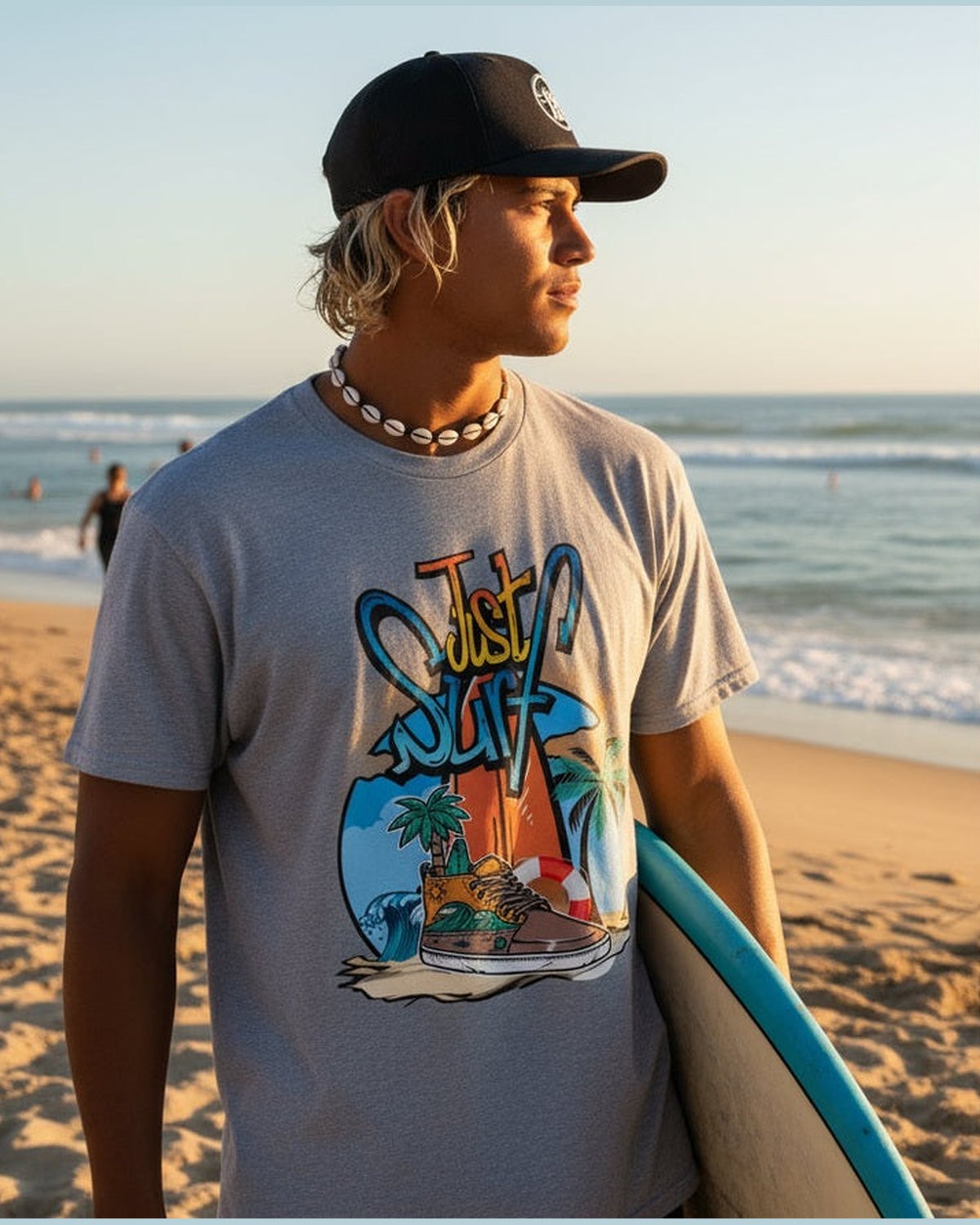 A surfer wearing a grey t-shirt with 'Just Surf' graphic design featuring a shoe, palm trees, and ocean at a beach setting 