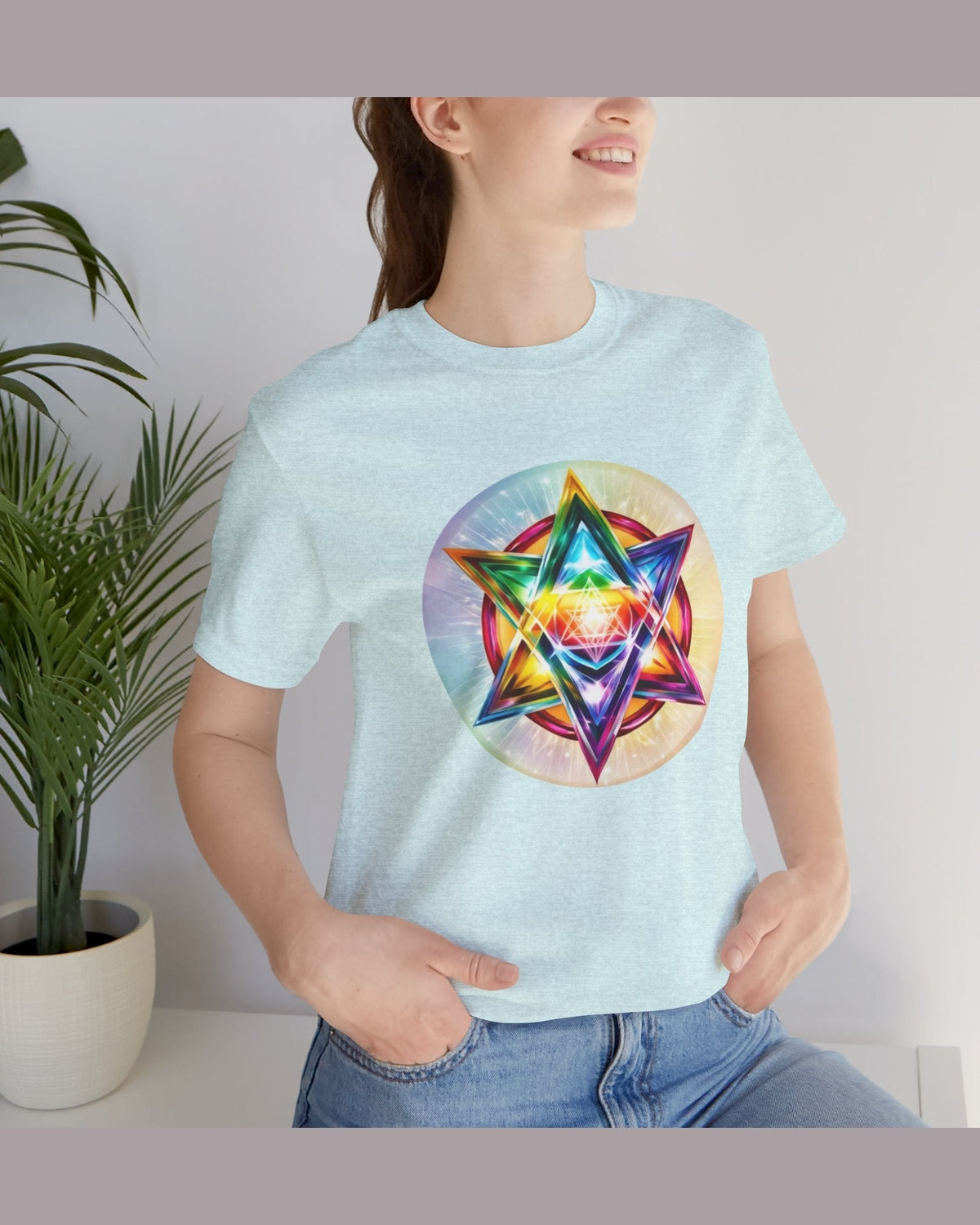 Person wearing a light blue t-shirt with a colorful Merkaba design, standing indoors next to a plant.