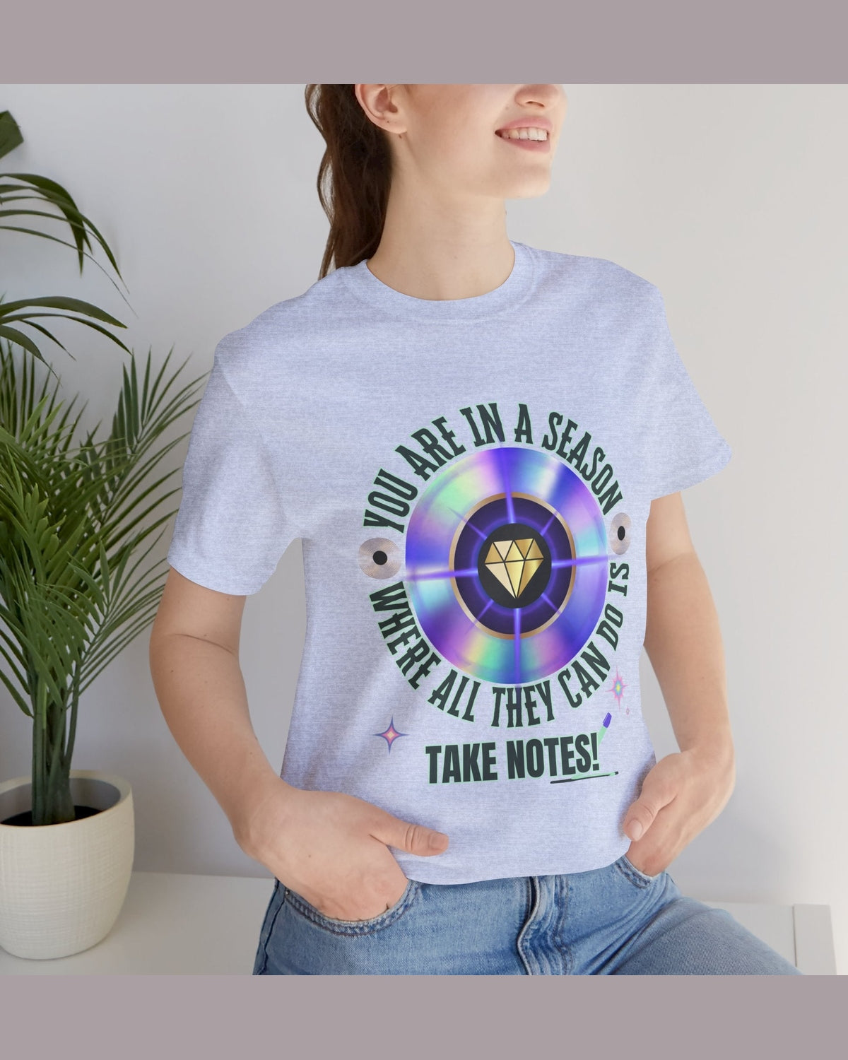 Person wearing a light blue t-shirt with a colorful graphic and text You are in a season where all they can do is take notes, standing indoors next to a plant