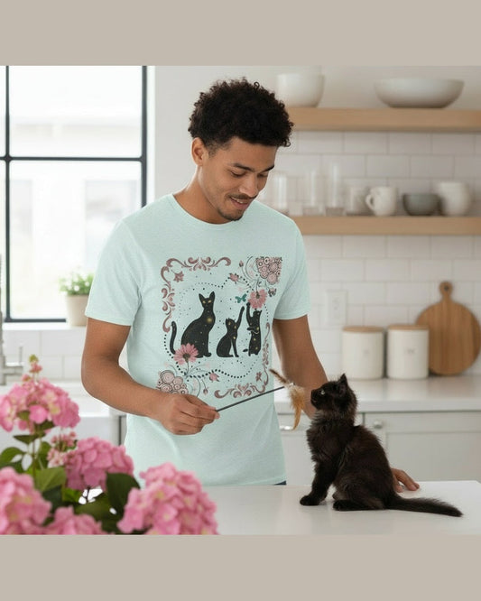 Man wearing a light blue t-shirt with cats and pink floral design in a kitchen with a black kitten on a counter 