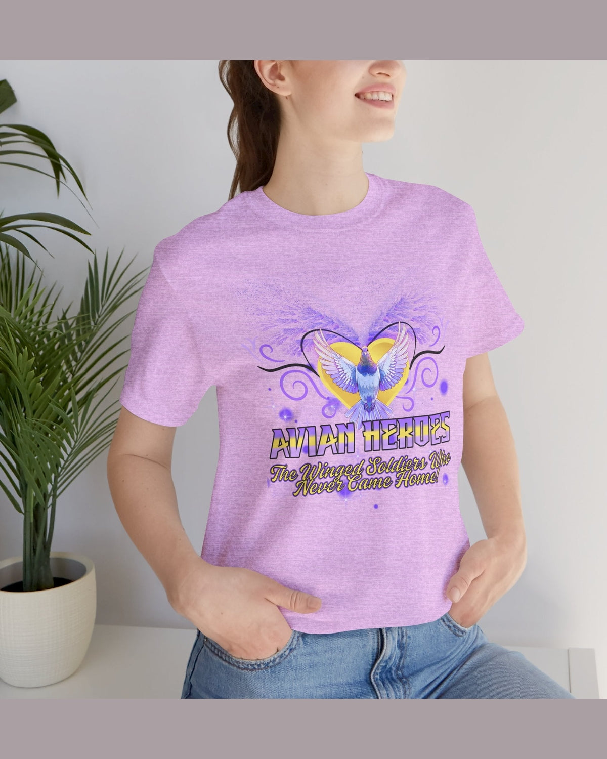 Person wearing a purple t-shirt with a tribute pigeon design and text Avian Heroes, standing indoors next to a plant.