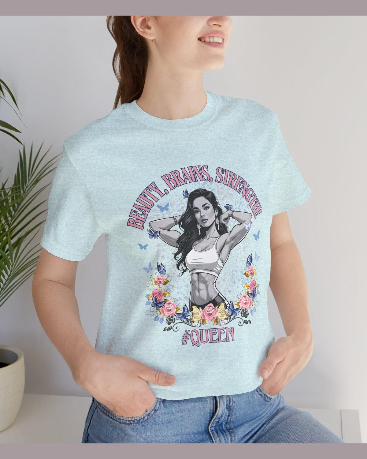 Person wearing a light blue t-shirt with a graphic design of a fitness woman, flowers, butterflies and text 'Beauty, Brains, Strength #Queen' standing indoors.