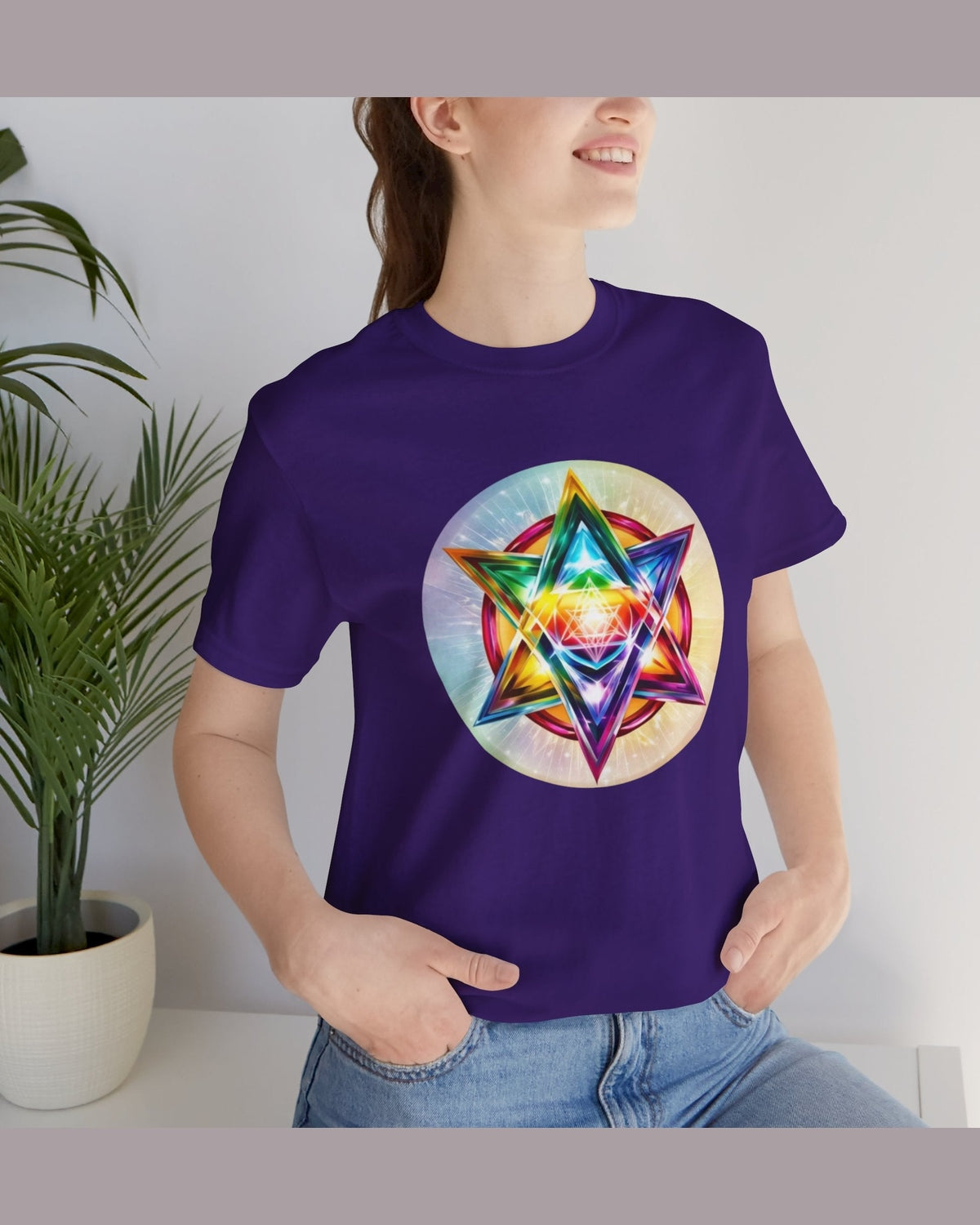Person wearing a purple t-shirt with a colorful Merkaba design, standing indoors next to a plant.