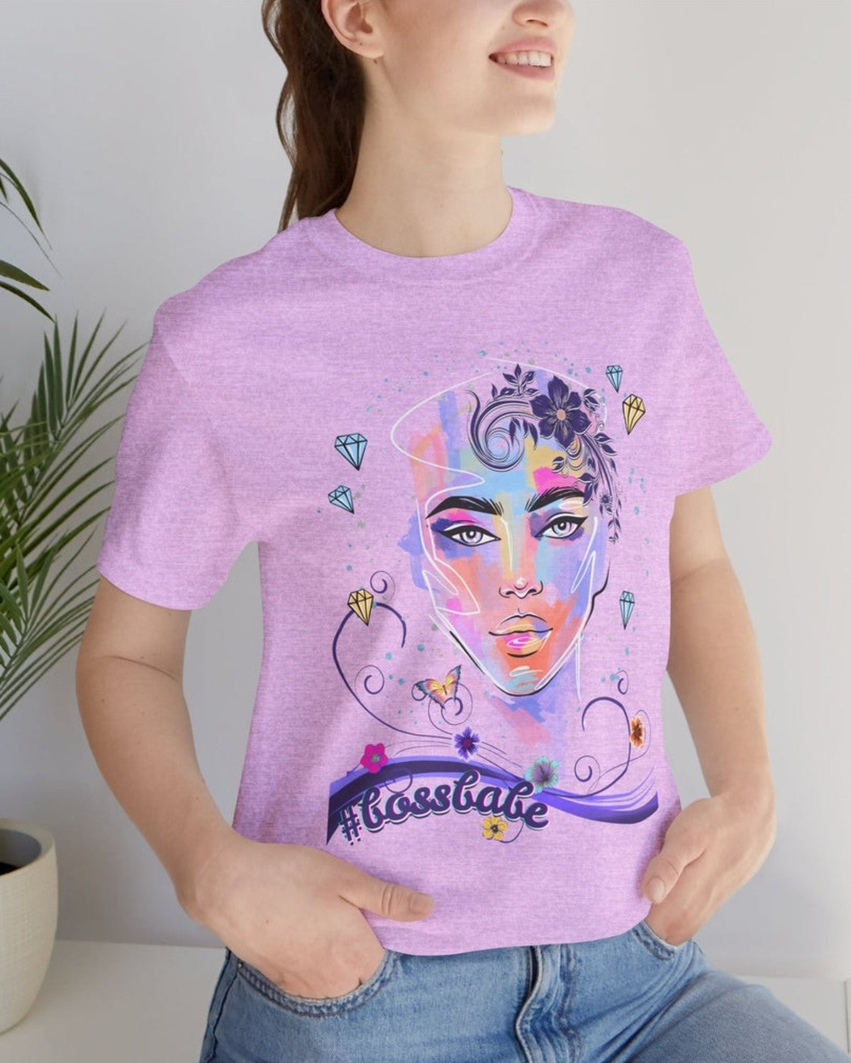 Person wearing a purple t-shirt with a colorful graphic design of a female face, diamonds and flowers and a text #bossbabe, sitting indoors next to a plant.
