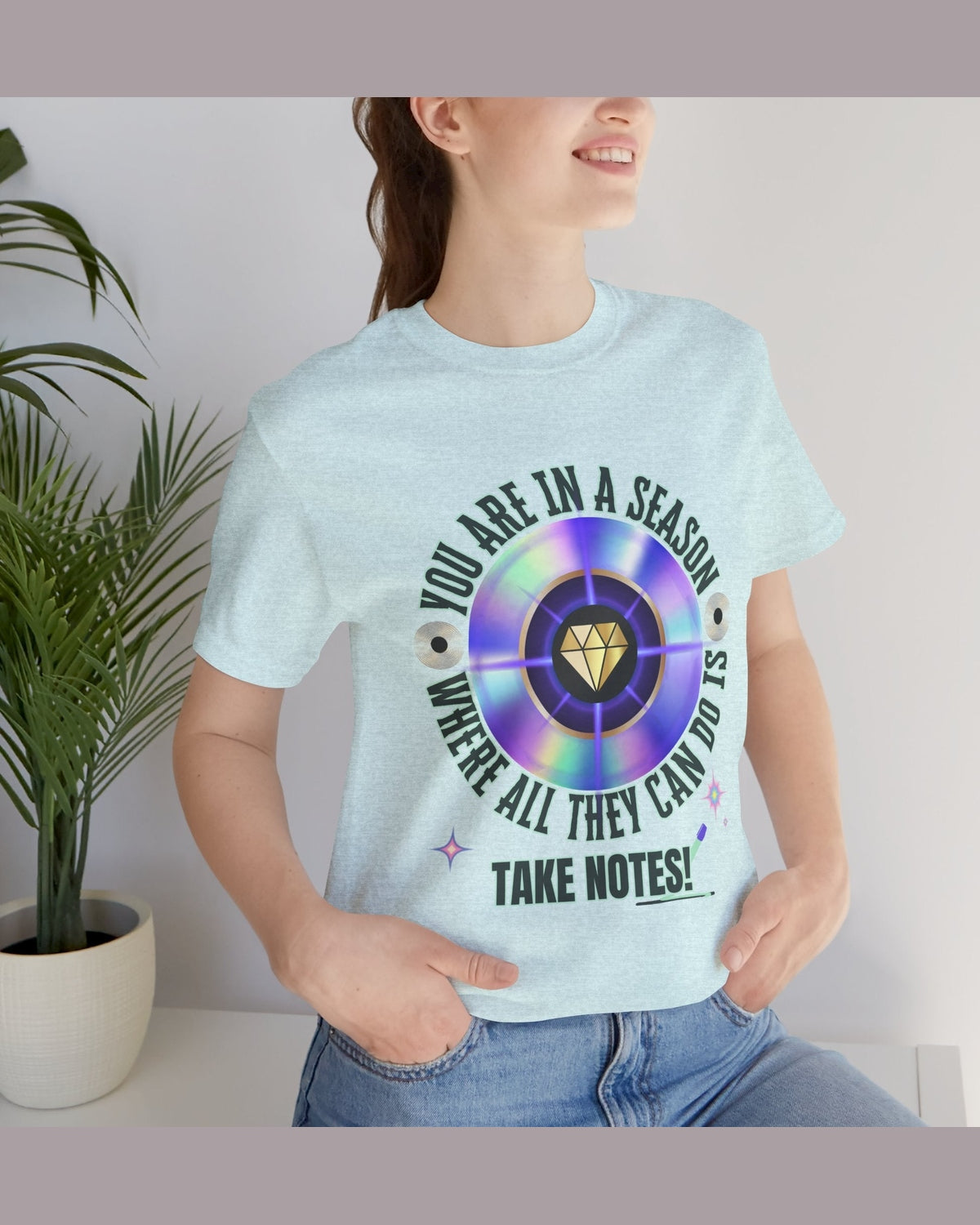Person wearing a light blue t-shirt with a graphic design and text 'You are in a season where all they can do is take notes', standing indoors next to a plant.