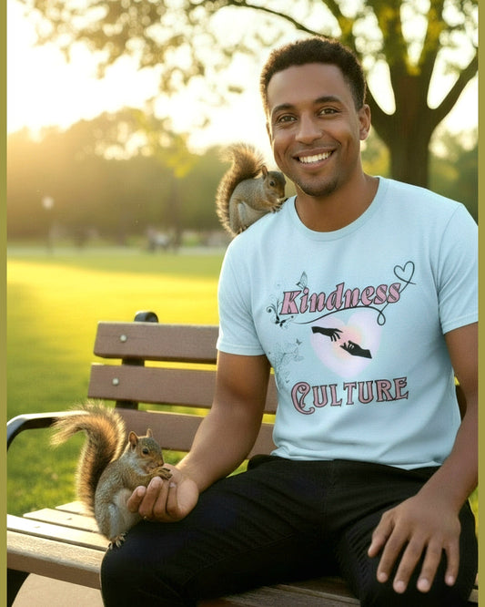 Man wearing Light blue t-shirt with 'Kindness Culture' text and hearr graphic sitting in a park feeding the squirrels 