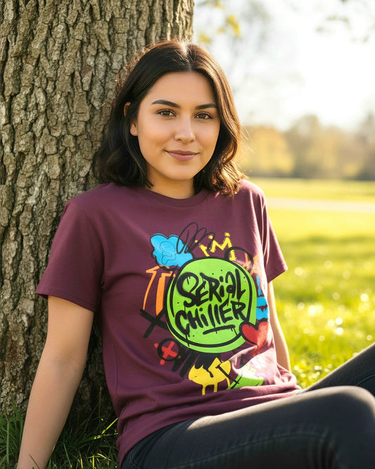 Woman wearing Maroon t-shirt with colorful 'Serial Chiller' grafitti graphic design sitting against a tree