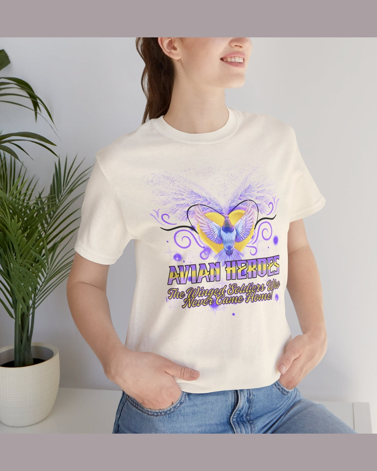Person wearing a soft yellow t-shirt with a pigeon and golden heart design and text Avian Heroes, standing indoors next to a plant.