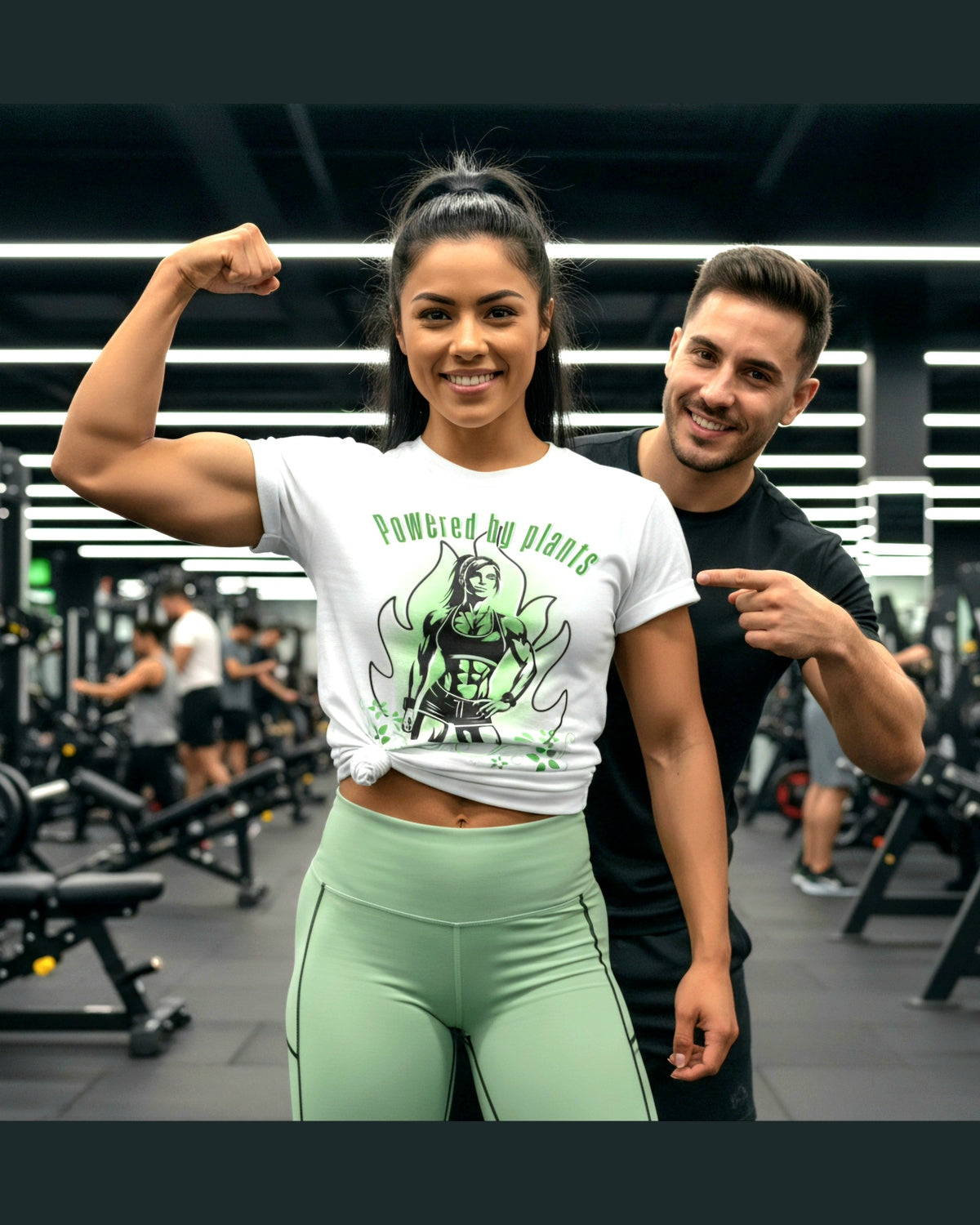 Woman flexing her arm in a gym wearing a 'Powered by plants' t-shirt, with a man pointing at her.